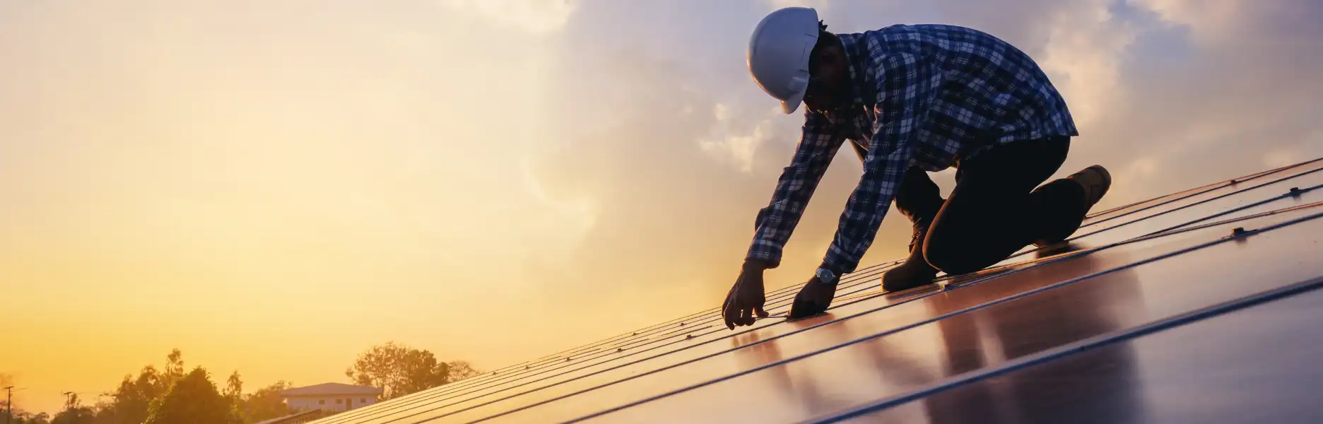 A technician in a hard hat and plaid shirt installing solar panels on a rooftop at sunset, with the sky fading from warm orange to deep blue.